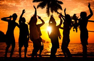 A tropical view beside water with six people, comprising of males and females, silhouetted by a setting sun. The people have stances, as if dancing, with their hands in the air. Directly behind them is the silhouette of a palm tree and further away are outcroppings of land.